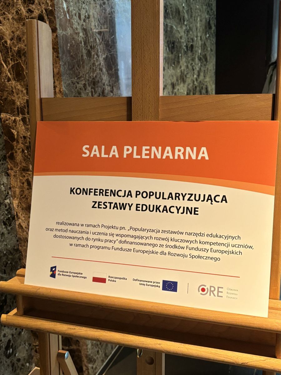 Na zdjęciu znajduje się plansza stojąca na sztaludze z napisem Sala Plenarna: Konferencja popularyzująca zestawy edukacyjne razem z informacją napisana drobnym drukiem że jest realizowana w ramach projektu unijnego dofinansowanego ze środków Funduszy Europejskich w ramach programu Fundusze Europejskie dla Rozwoju Społecznego. Plansza jest utrzymana w kolorach ciepłego pomarańczu oraz brązu