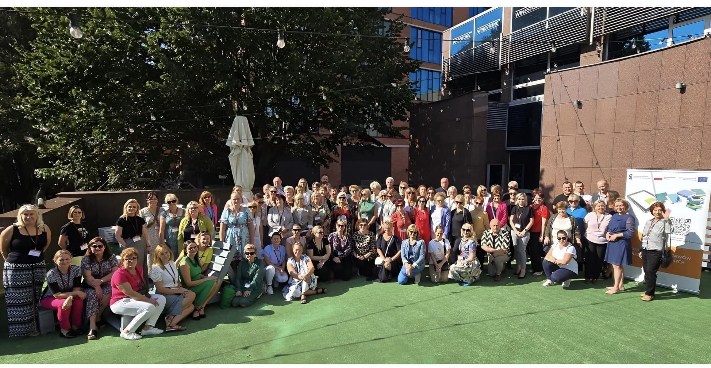 Zdjęcie przedstawia grupę wszystkich uczestników konferencji: organizatorów, doradców metodycznych, nauczycieli oraz konsultantów którzy są ustawieni na tarasie hotelu Mercure w Gdańsku do zdjęcia grupowego. Osoby są uśmiechnięte, atmosfera jest wyraźnie pozytywna, świeci słońce, z tyłu widoczne są zielone drzewa 