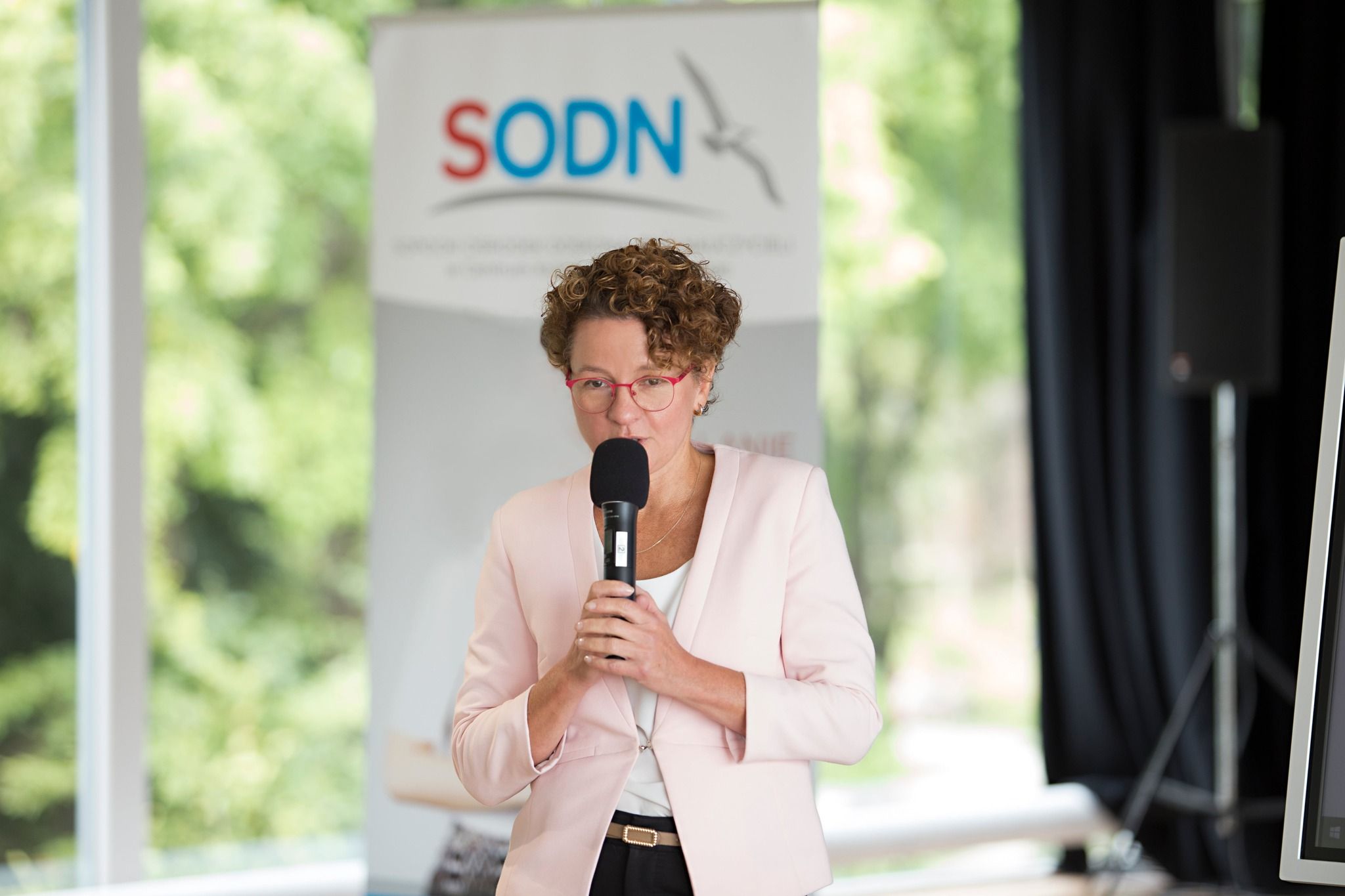 Prezydentka Miasta Sopotu - Pani Magdalena Czarzyńska-Jachim stoi na tle roll-up z logo SODN. Trzyma w ręku mikrofon. Wita zgormadzonych uczestników konferencji.